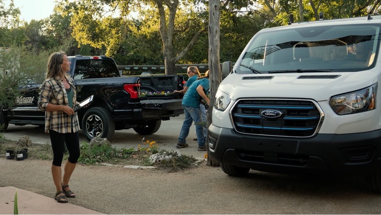 On the right, a Ford E-Transit. On the left, a Ford F-150. Two Ford Pro customers, a woman and man, are working in between them.