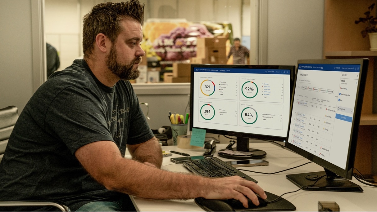 Homme assis à un pupitre devant deux écrans d’ordinateur avec une main sur une souris informatique. En arrière-plan, il y a un employé, des boîtes et une fourgonnette.