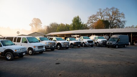 A mixed fleet of vans, trucks, and haulers in front of a motor pool structure.