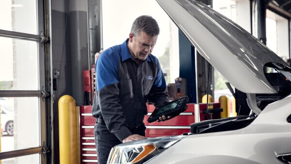 A service technician holding a tablet, looking under the hood of a vehicle.