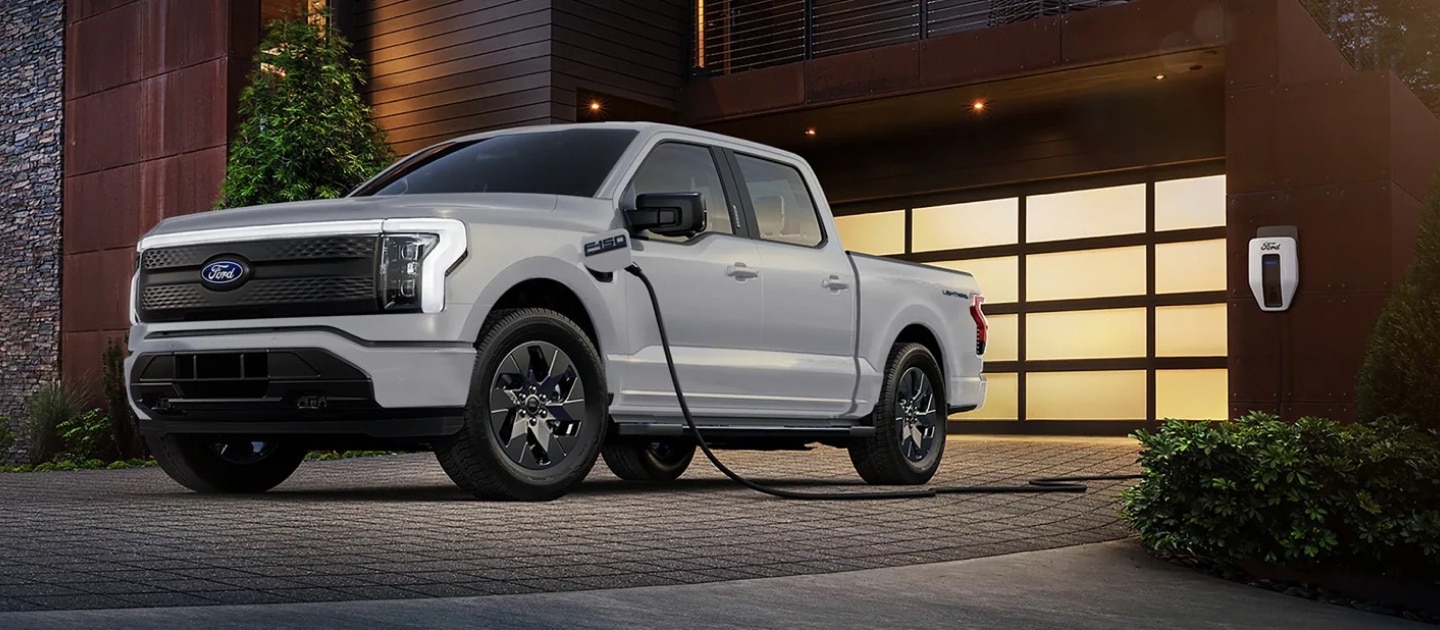 A grey 2024 Ford F-150 Lightning pickup truck parked in the driveway of a home in the woods