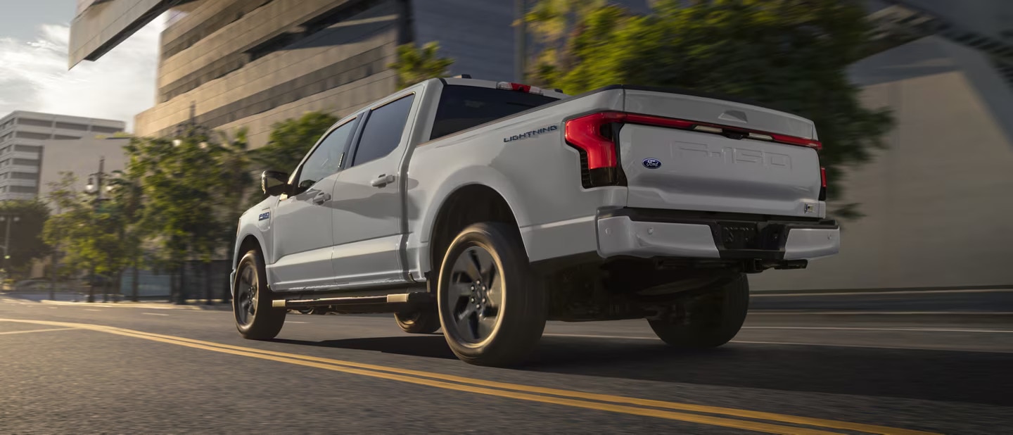 A white 2024 Ford F-150 Lightning pickup truck driving near a large glass office building in the late afternoon