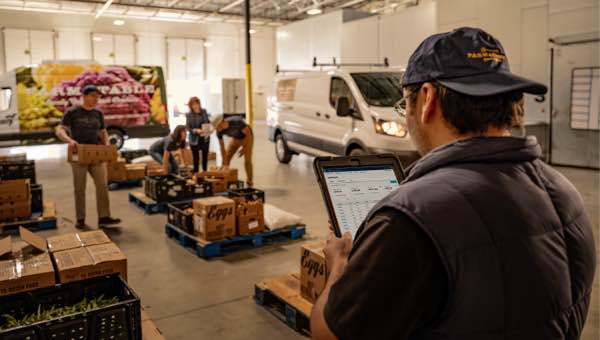 Man on tablet looking at Ford Pro™ Telematics software as workers move boxes in a warehouse