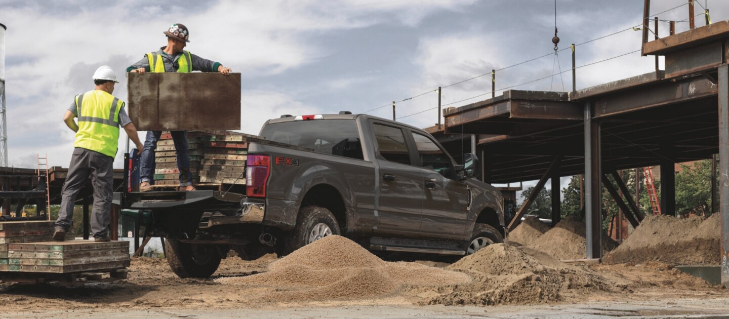 Camioneta Ford F-250 gris en obra de construcción, con dos trabajadores manipulando materiales pesados en la batea.