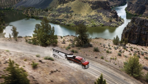 Camioneta Ford Super Duty roja arrastrando un remolque por un camino de tierra en un paisaje montañoso con un río.