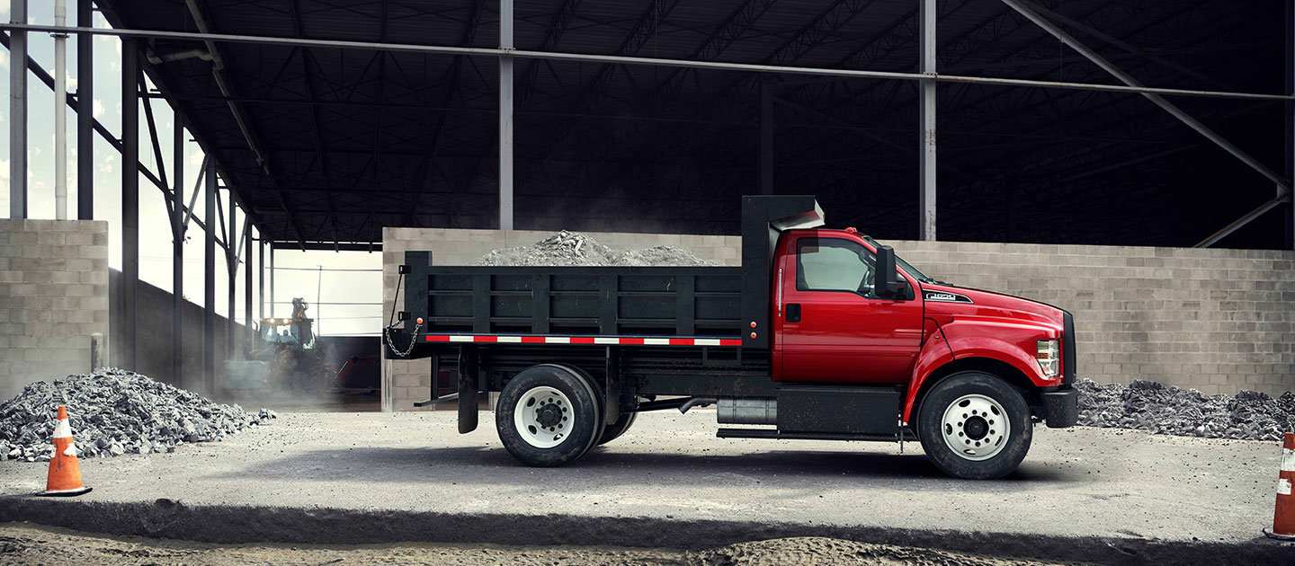 Un camión Ford de cabina roja y caja negra cargada de grava está estacionado de perfil frente a una gran estructura industrial de acero gris y muros de bloques, con pilas de escombros y un cono naranja en primer plano.