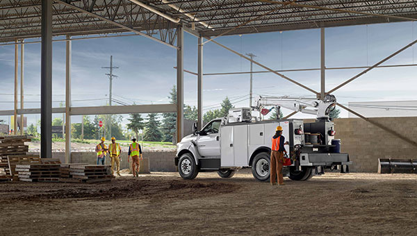 Un camión de servicio Ford blanco equipado con una grúa trasera está estacionado bajo la estructura de acero gris de una nave en construcción, rodeado de trabajadores con chalecos reflectantes y pilas de palés de madera sobre un suelo de tierra.