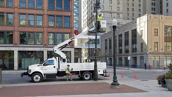 Un trabajador en una Plataforma elevadora Versalift blanca repara una luminaria junto a un camión Ford blanco, situado frente a edificios urbanos de ladrillo rojo y piedra clara con conos naranjas en el pavimento.