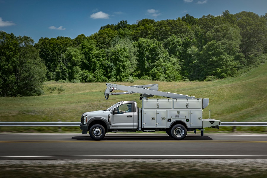 Ford Super Duty Chasis blanca con una grúa o pluma en la parte trasera se desplaza por una carretera junto a un paisaje de colinas verdes y árboles.