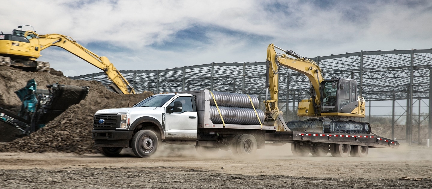 Camioneta Ford Super Duty blanca en obra de construcción, arrastrando un remolque con una excavadora y transportando materiales.