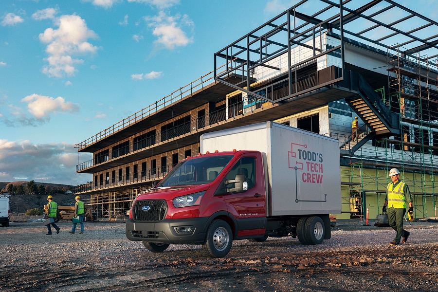 Ford Transit Chasis roja con carrocería de servicio estacionada a lado de una estructura metálica en construcción, con trabajadores.