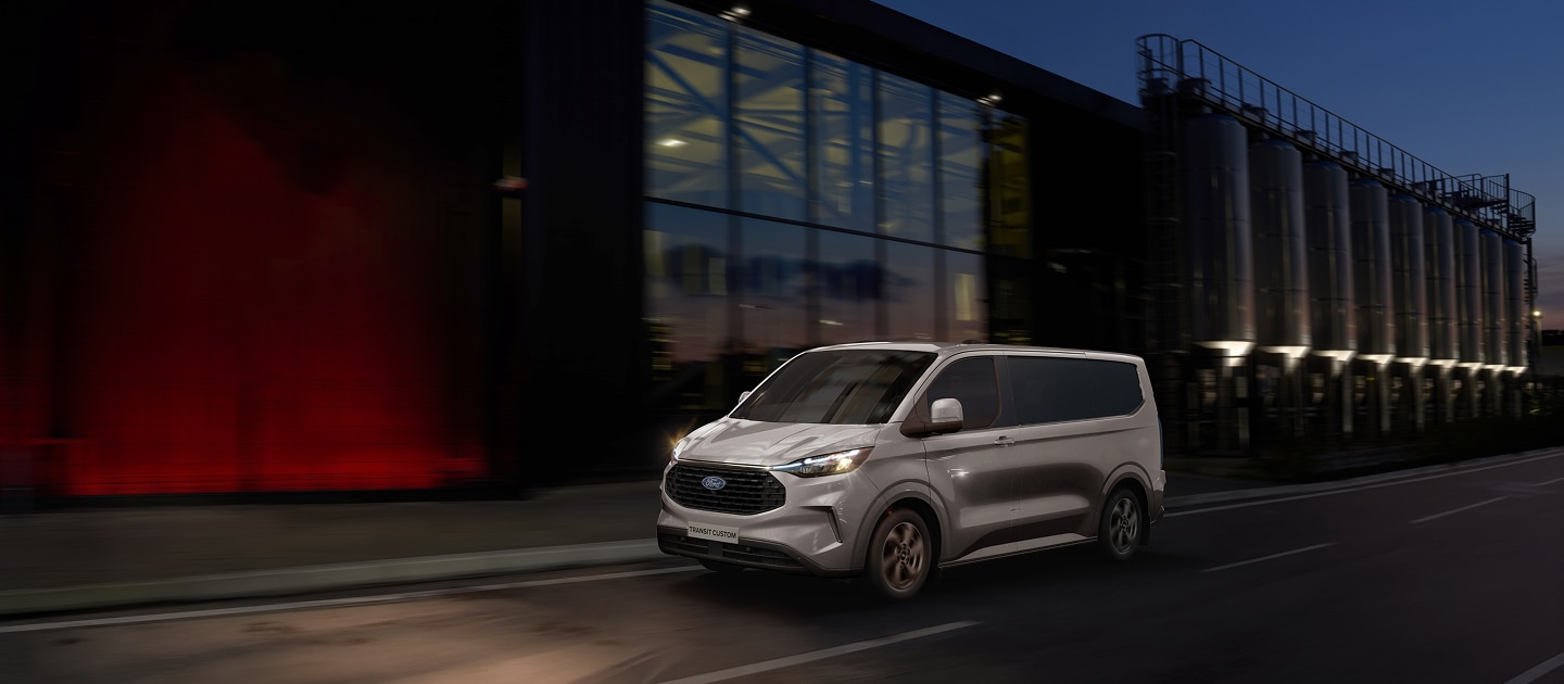 Ford Transit Custom 2026 plata en movimiento al anochecer con faros encendidos.
