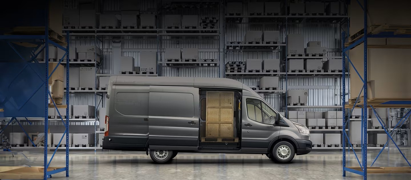 Ford Transit Van color gris oscuro con la puerta lateral abierta, mostrando cajas de madera en su interior, estacionada dentro de un almacén con estanterías y mercancía.