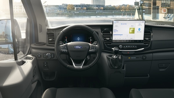 Dashboard de una Ford Transit Van mostrando el volante, el clúster digital y una gran pantalla táctil central, con vista a un paisaje urbano y acuático a través del parabrisas.