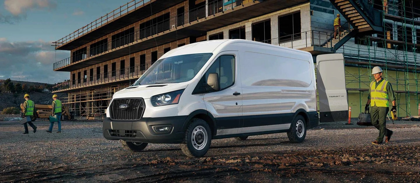 Ford Transit Van blanca estacionada en un sitio de construcción con la puerta trasera abierta, mientras un trabajador con casco y chaleco de seguridad camina cerca.