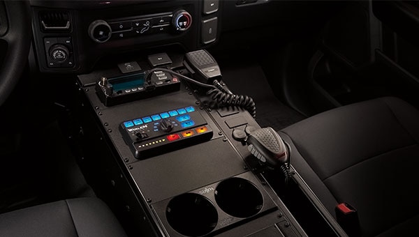 Vista en primer plano de la consola central de una Ford Police Interceptor, mostrando equipos policiales como una radio, un panel de control Whelen y un micrófono, instalados en el espacio de accesorización modular del vehículo.