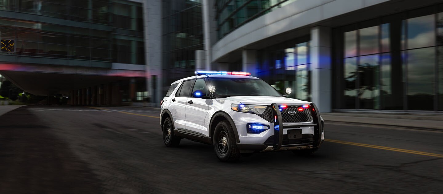 Police Interceptor Híbrida con luces de emergencia azules y rojas encendidas en el techo y en la parte frontal cruzando una calle rodeada de edificios a alta velocidad.