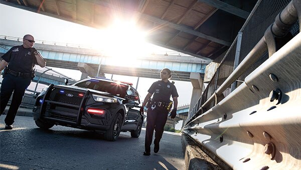 Dos oficiales de policía caminan junto a una patrulla Ford Police Interceptor Híbrida negra estacionada bajo un puente elevado, con la luz del sol brillando intensamente al fondo.