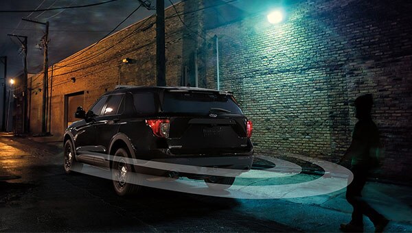 Vista trasera de un Ford Police Interceptor negro estacionado de noche en un callejón iluminado por farolas, con un sistema de alerta perimetral visualizado  alrededor del vehículo que detecta a una persona caminando en el lado derecho de la imagen.