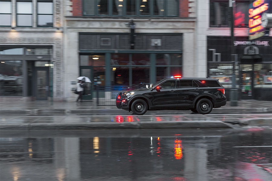 Vista lateral de Ford Police Interceptor Utility negra en una calle urbana durante el día, cerca de un edificio moderno de cristal. El vehículo tiene una pequeña luz de emergencia roja en el techo.