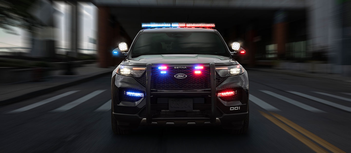 Vista frontal de un Ford Police Interceptor Utility negro en movimiento rápido, con sus luces de emergencia rojas y azules encendidas en el techo y la parrilla, y una defensa delantera, sobre una calle urbana con el fondo borroso.