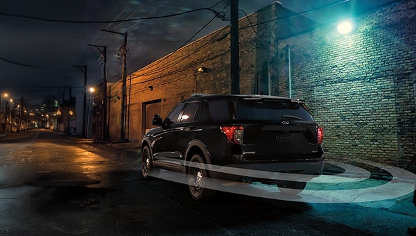 Vista trasera de un Ford Police Interceptor Utility negro estacionado de noche en un callejón iluminado por farolas, con un sistema de alerta perimetral visualizado alrededor del vehículo que detecta a una persona caminando en el lado derecho de la imagen