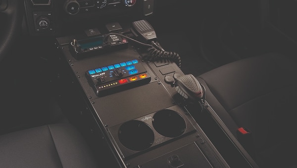 Vista en primer plano de la consola central de una Ford Explorer Police Interceptor, mostrando equipos policiales como una radio, un panel de control Whelen y un micrófono, instalados en el espacio de accesorización modular del vehículo.