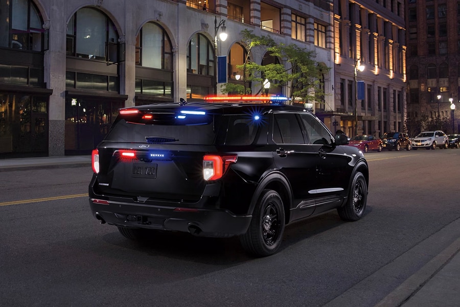 Vista trasera de Ford Police Interceptor Utility negra en una calle urbana de noche, con sus luces de emergencia rojas y azules encendidas en el techo y la parte trasera.