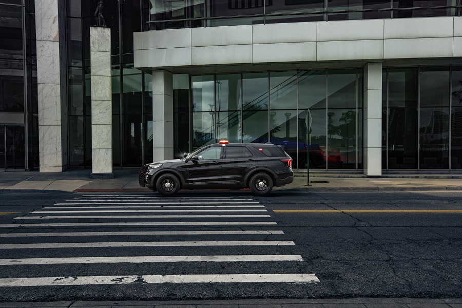 Vista lateral de Ford Police Interceptor Utility negra en una calle urbana durante el día, cerca de un paso de peatones y un edificio moderno de cristal. El vehículo tiene una pequeña luz de emergencia roja en el techo.