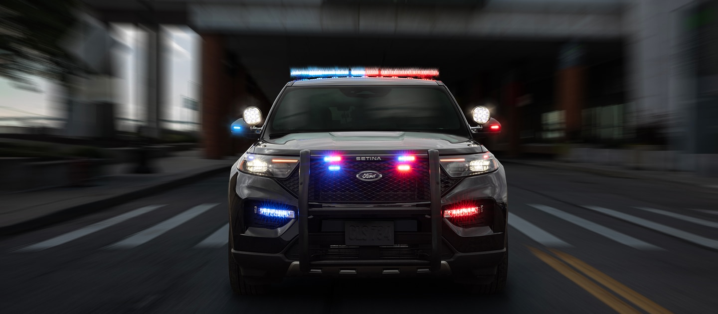 Vista frontal de un Ford Police Interceptor Utility negro en movimiento rápido, con sus luces de emergencia rojas y azules encendidas en el techo y la parrilla, y una defensa delantera, sobre una calle urbana con el fondo borroso.