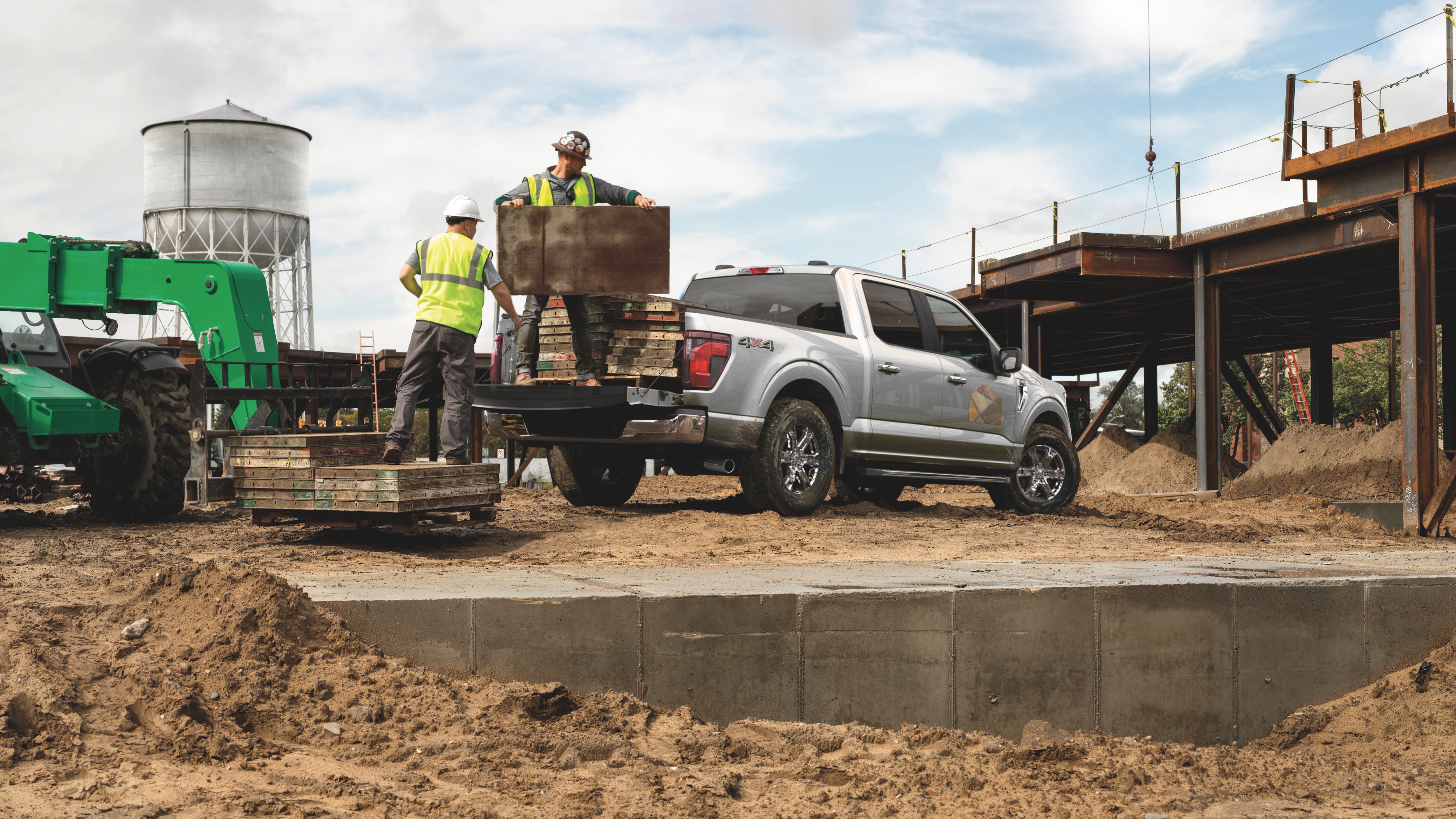 F-150 azul en obra de construcción, dos trabajadores cargando materiales en la caja, con maquinaria pesada al fondo.