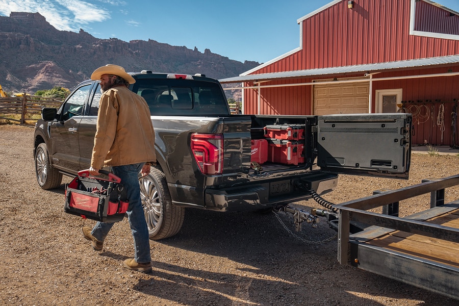 Un hombre con sombrero transporta herramientas mientras la batea multiacceso de Ford Lobo 2025 está abierta lateralmente, mostrando cajas de herramientas en su interior y un remolque enganchado.