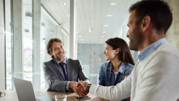Tres personas en una oficina moderna participando en una reunión de negocios, con dos individuos estrechando la mano a través de una mesa.