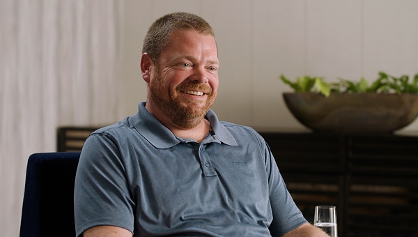 Photograph portrays smiling middle-aged man seated indoors, during a casual interview. Dark furniture, curtains, and leafy bowl-shaped planter form a relaxed background.