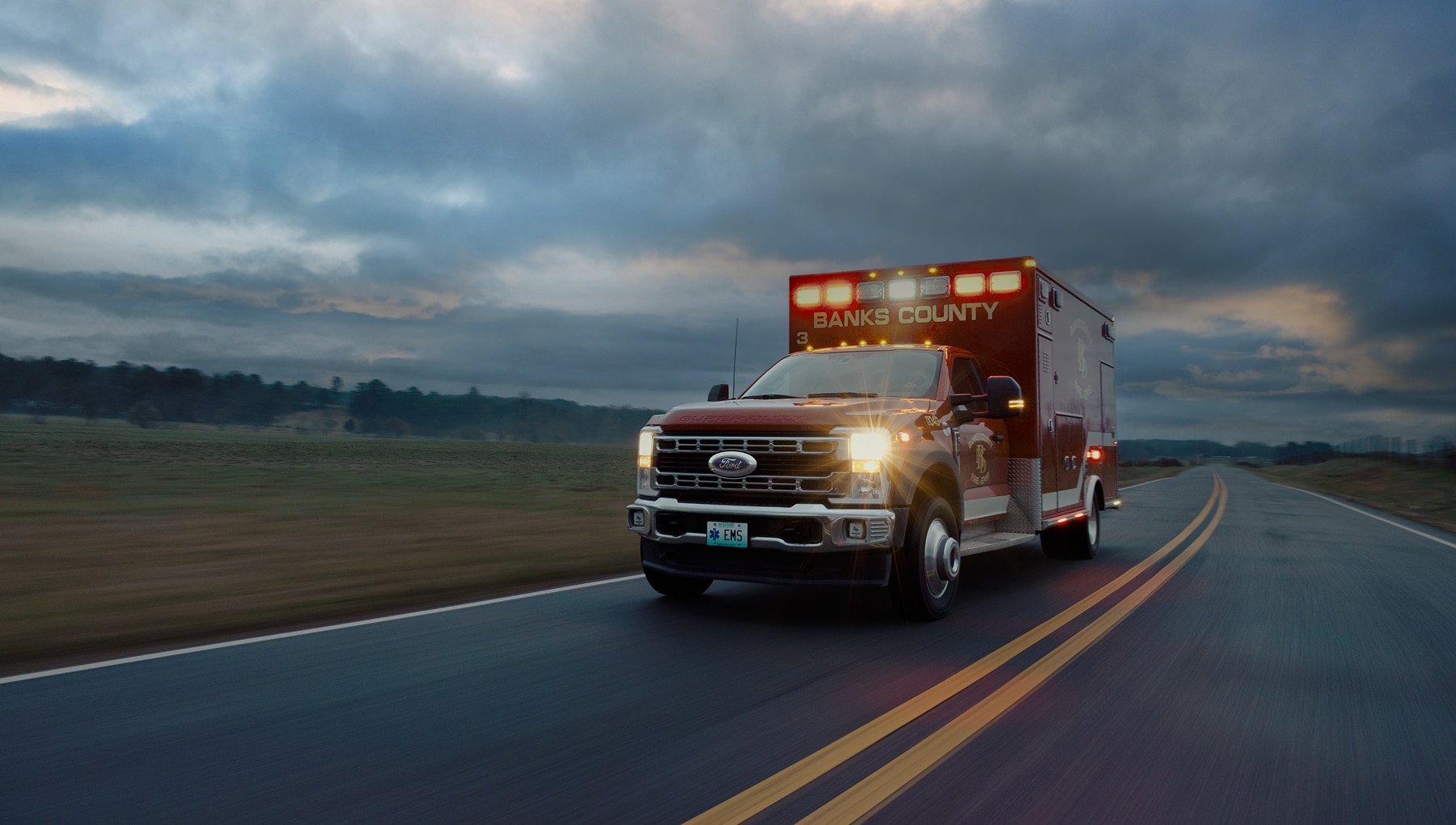 Red Ford Super Duty ambulance with flashing emergency lights driving on a rural road at dusk.