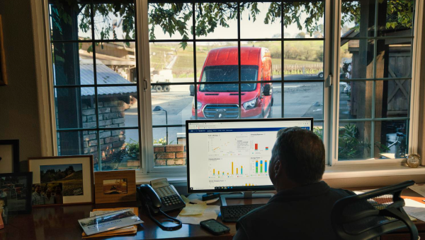 A man working on a computer at a desk facing a window with a 2023 Ford Transit van parked outside.