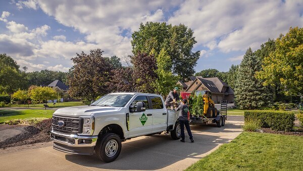 A white Super Duty truck is parked in a driveway. Landscapers unload towed equipment.