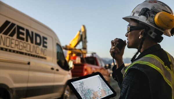 Construction worker in safety gear using a tablet and radio near utility vans and heavy equipment at a worksite.