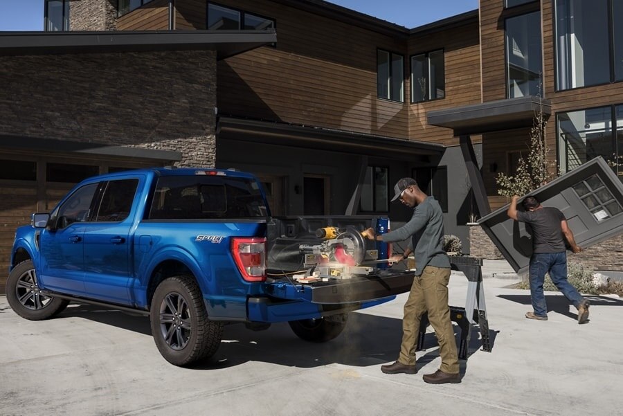 A man cutting wood with a power saw on the tailgate of a 2023 Ford F-150® XLT SuperCrew® in Atlas Blue in front of a modern home