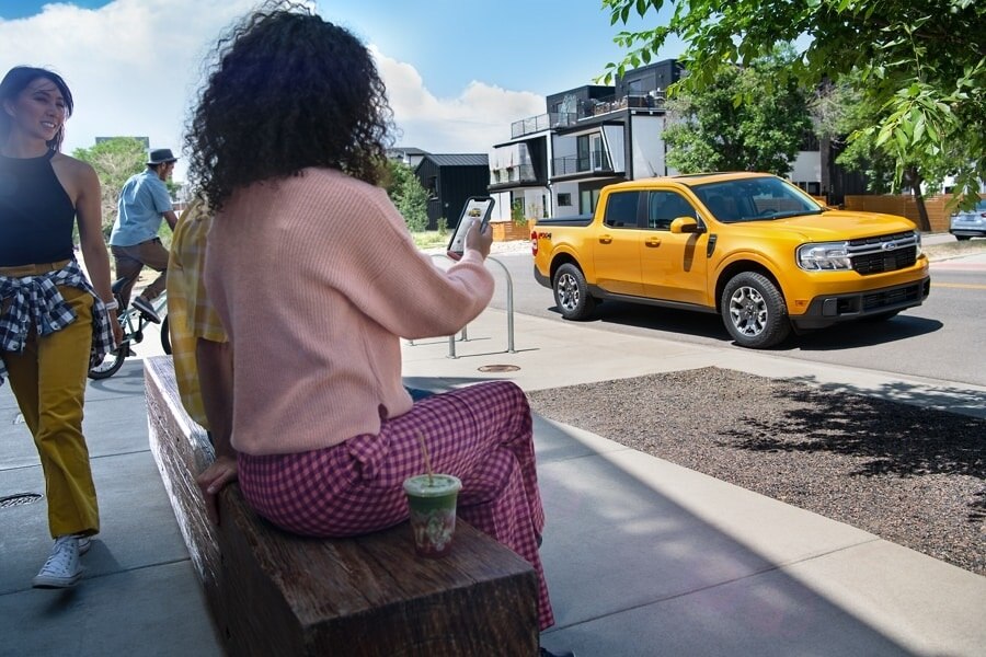Woman sitting on a bench looking at her phone with a 2023 Ford Cyber Orange Maverick® truck parked in the street