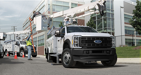 A white Ford Super Duty with a bucket truck upfit parked at a job site. A worker in a yellow vest hands an orange tube to a worker in the bucket.