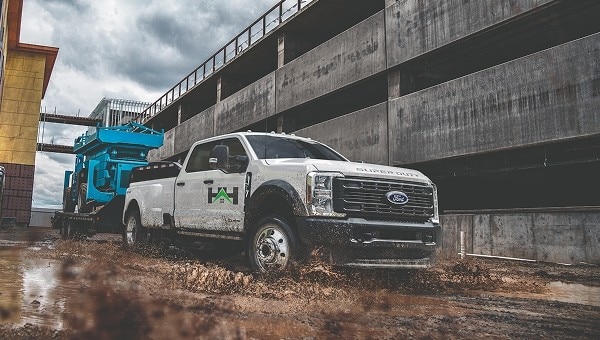 White Ford Super Duty truck towing a heavy machinery trailer through a muddy construction site.