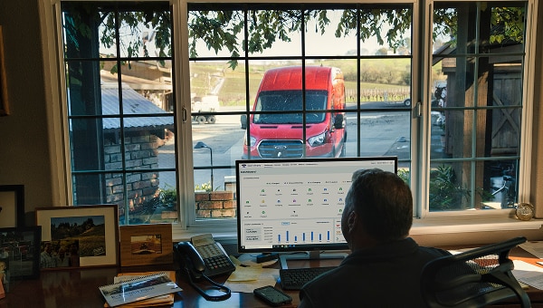 A person working on a computer at a desk facing a window with a 2023 Ford Transit van parked outside.