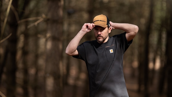 A man in a forest wearing a black Ford/Carhartt t-shirt and a branded beanie.